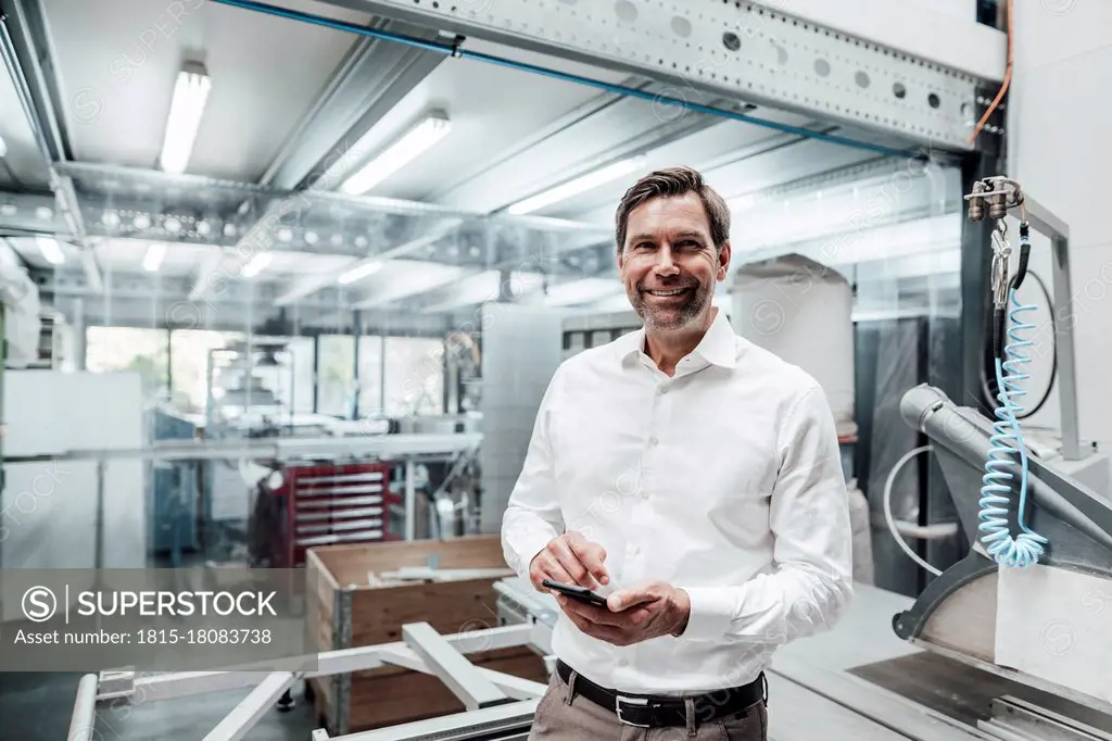 Smiling male engineer with smart phone standing by machinery in industry