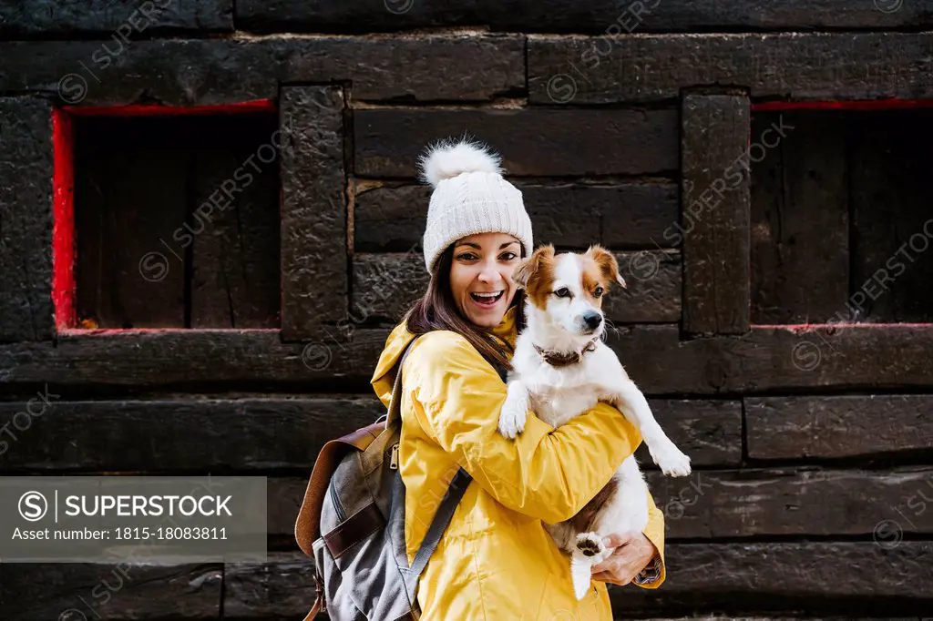 Portrait of smiling woman in yellow raincoat holding dog in front of log cabin