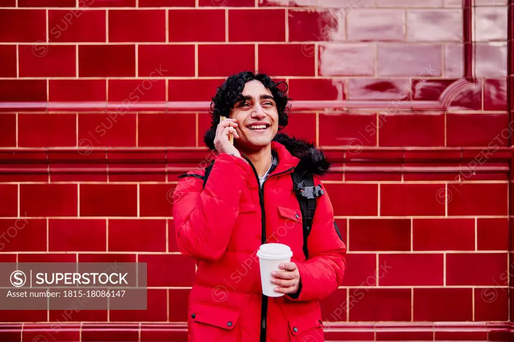 Smiling young man holding disposable coffee cup while talking on mobile phone against wall