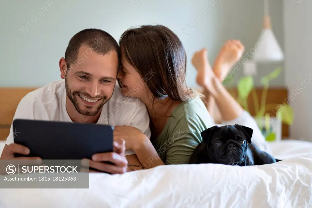 Woman embracing boyfriend looking at digital tablet while lying on bed in bedroom at home