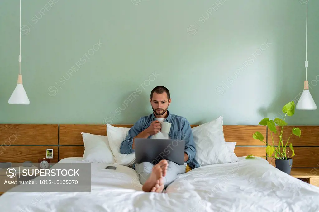 Young man having coffee using laptop while sitting on bed at home