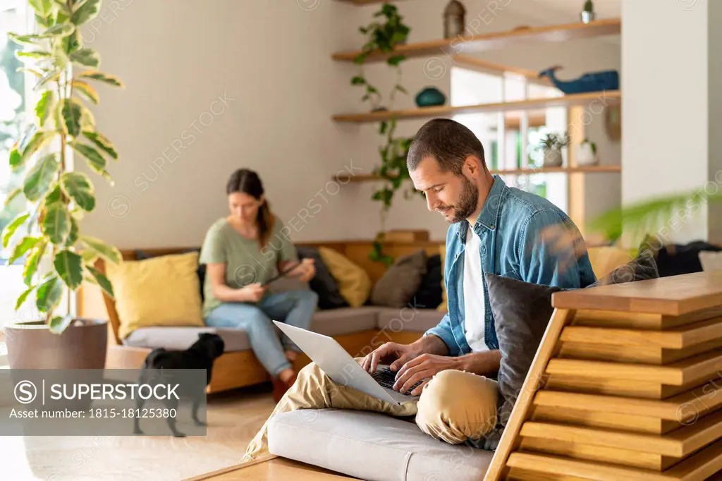 Young man working on laptop while wife looking at Pug dog in background at home