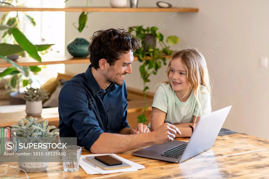 Smiling father and daughter looking at each other in front of laptop at home