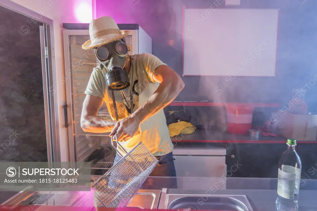 Mature man in gas mask preparing food in deep fryer at commercial kitchen