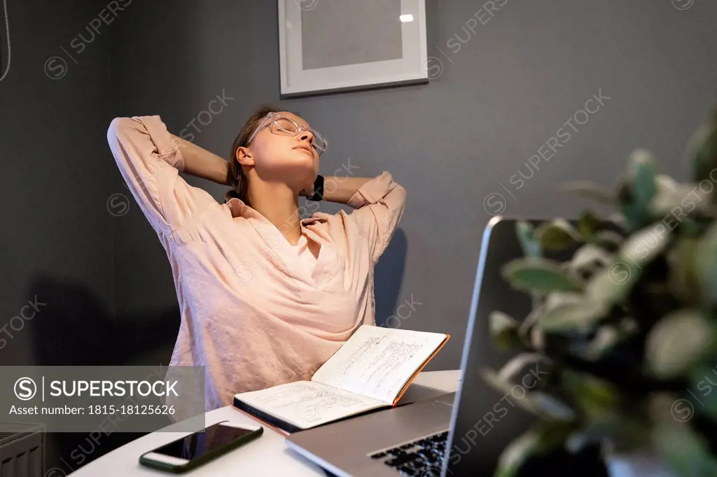 Tired woman sitting with hands behind head by laptop at home