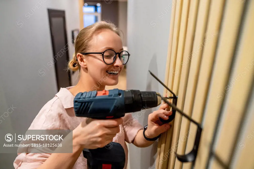 Smiling woman using drill machine while fixing hook on wall at home