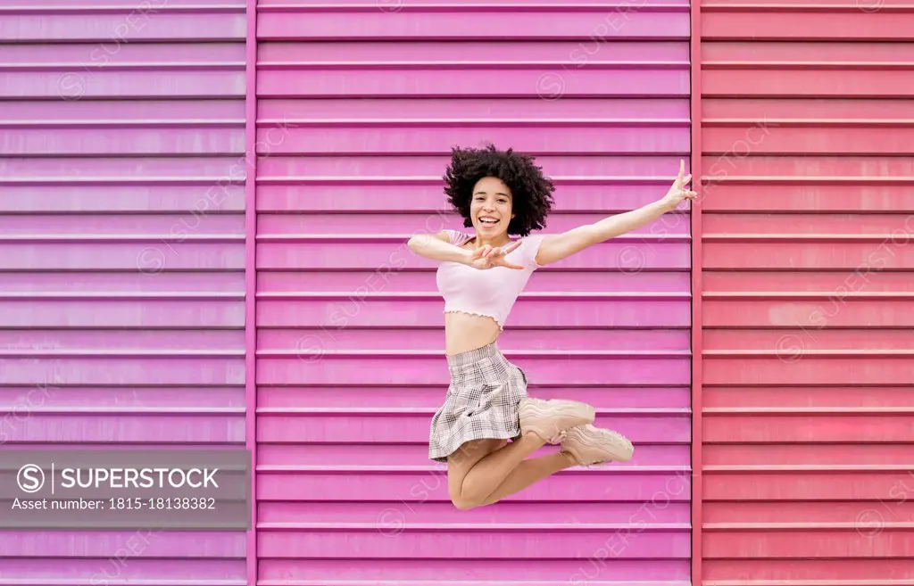 Carefree woman showing peace sign while jumping against multi colored wall