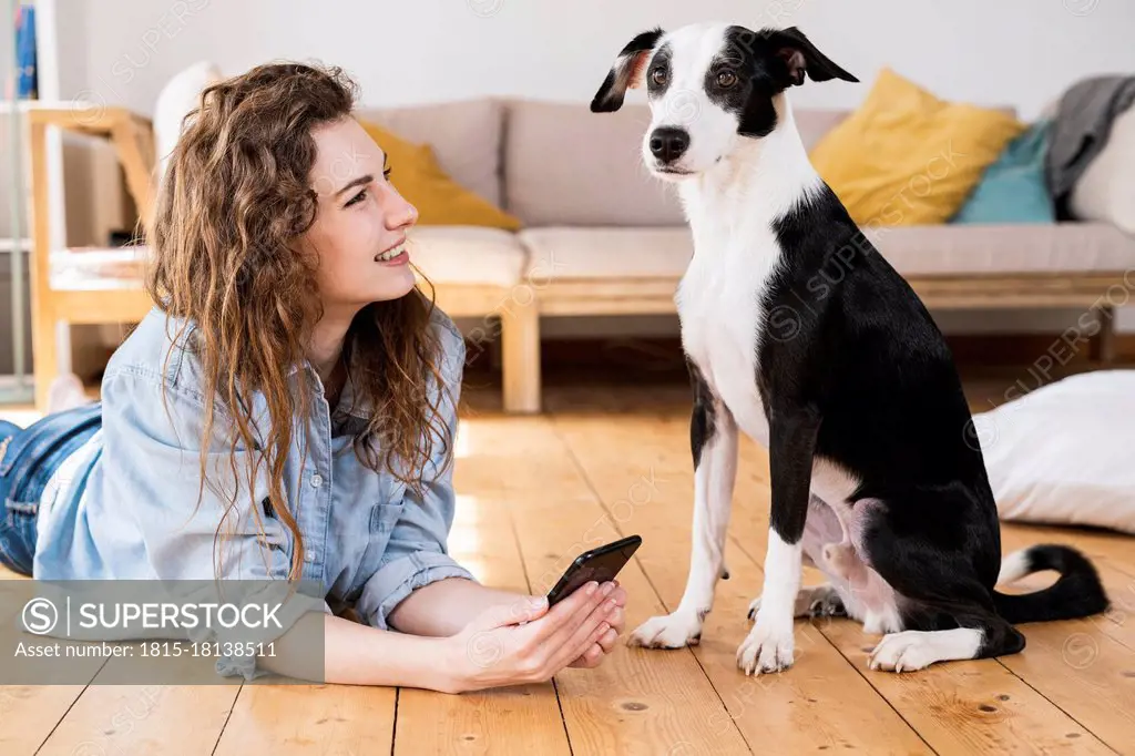 Beautiful woman with long hair holding smart phone while looking at dog in living room