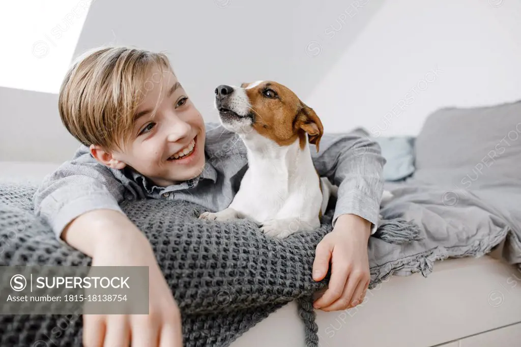 Smiling boy with arm around dog lying on bed at home