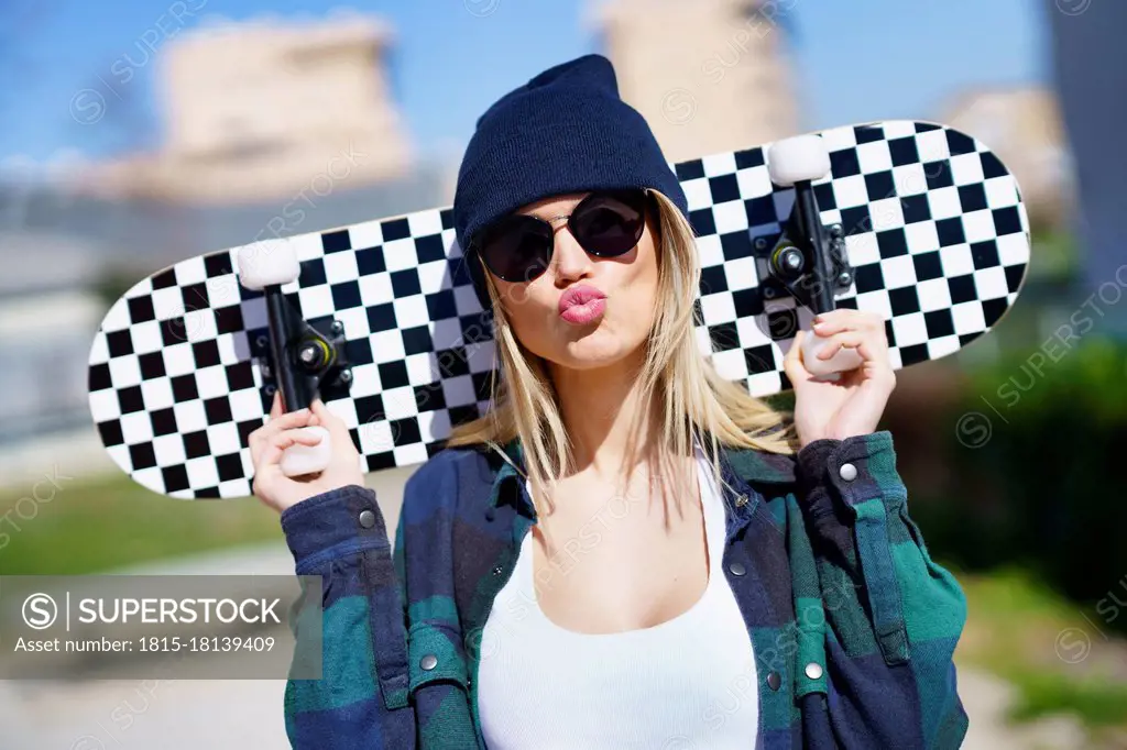 Young woman in knit hat puckering while carrying skateboard on shoulder during sunny day