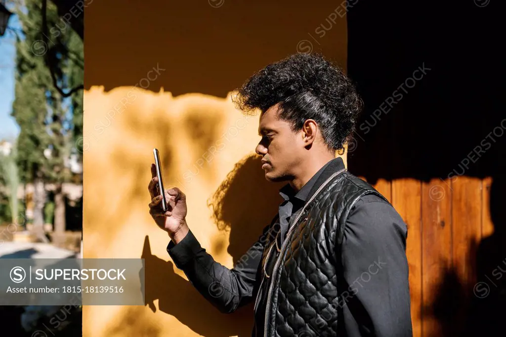 Mid adult man using phone while standing by wall on sunny day