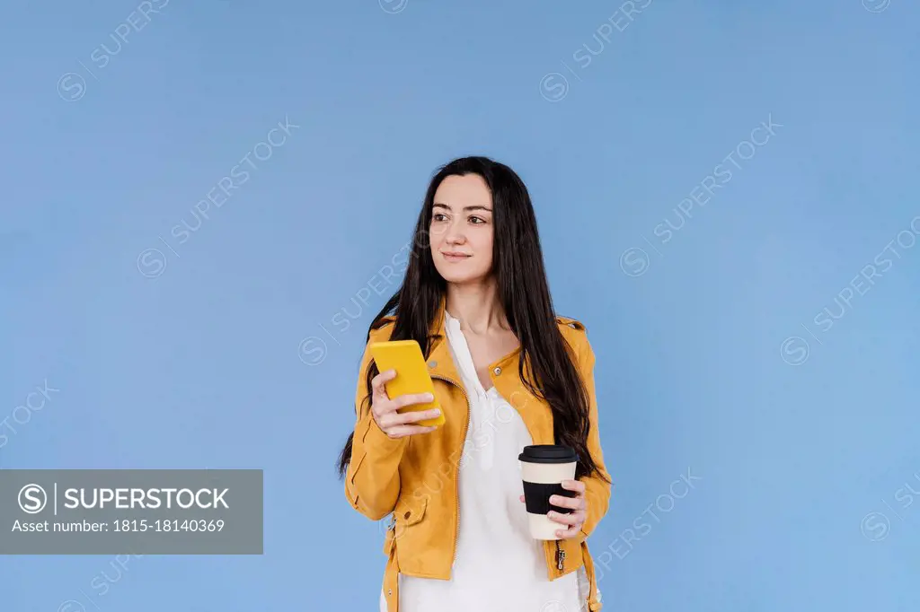 Smiling woman with mobile phone looking away while standing against blue background