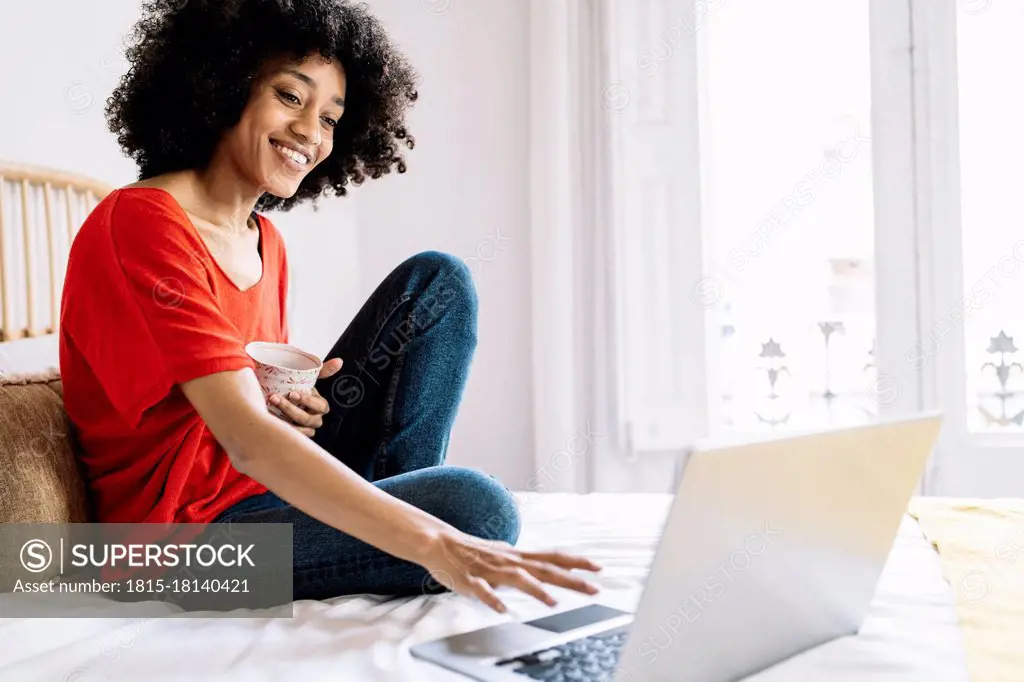 Young woman with mug using laptop while sitting on bed at home