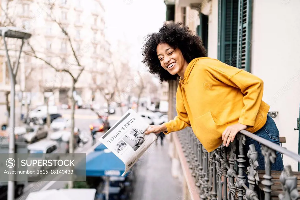 Cheerful young woman with newspaper leaning on railing in balcony