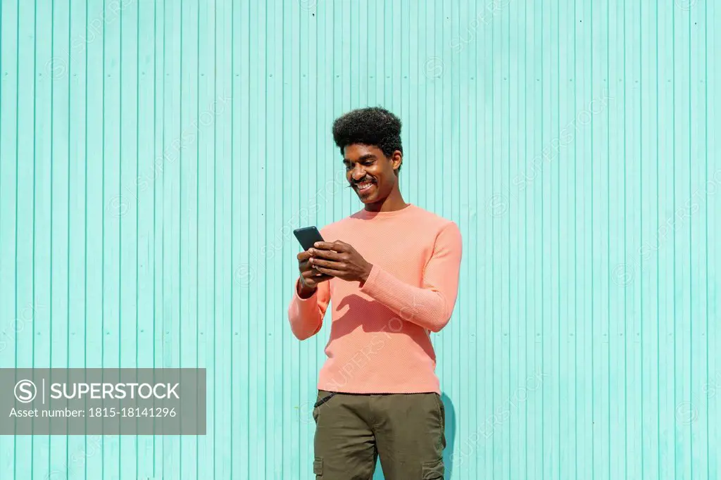 Smiling man using smart phone in front of turquoise wall during sunny day