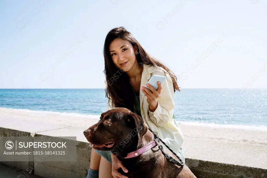 Smiling woman sitting with Labrador Retriever by sea