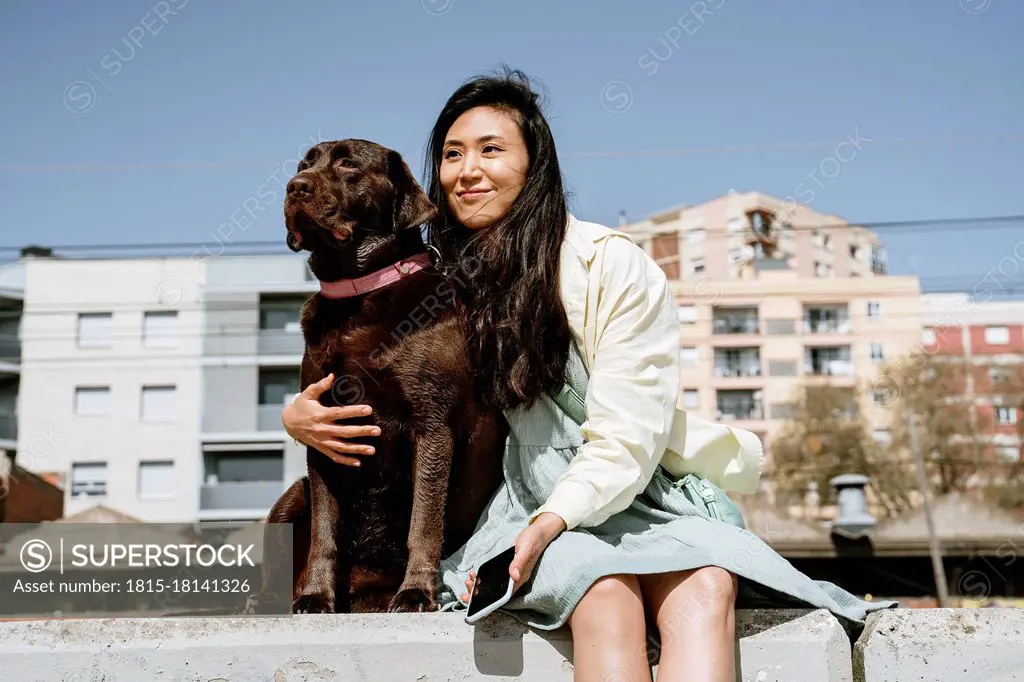 Smiling woman sitting with Chocolate Labrador on retaining wall in front of building