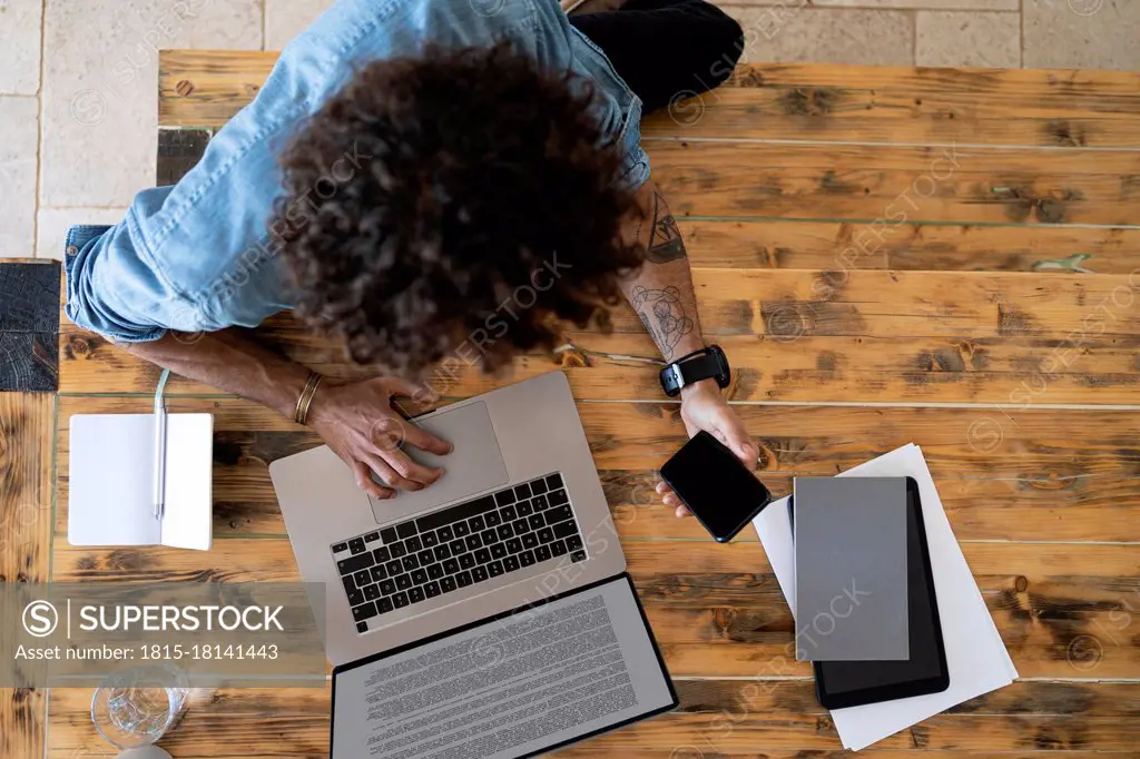 Male professional holding mobile phone while using laptop on table at home