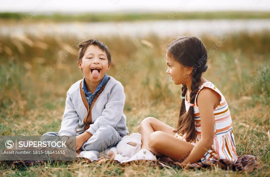 Girl looking at brother with down syndrome sticking out tongue while sitting on grass
