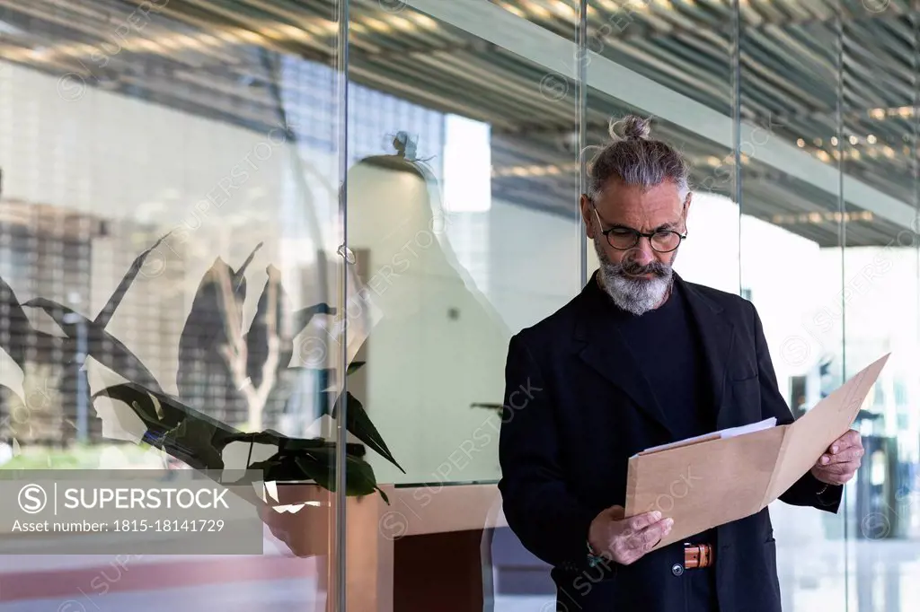 Male professional reading file while standing by glass wall