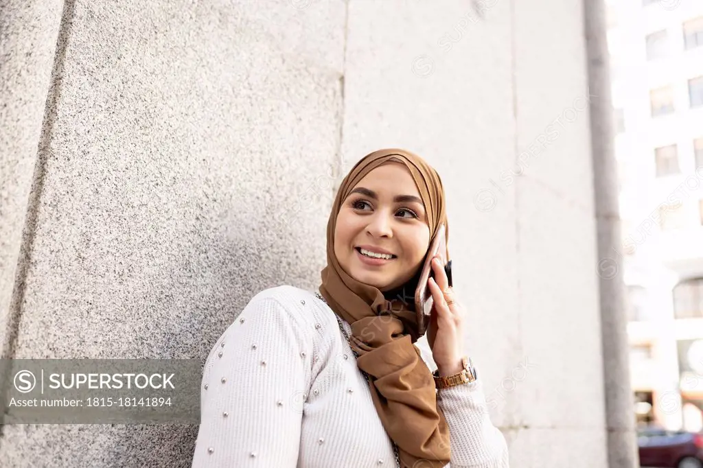 Young woman in hijab looking away while talking on smart phone by wall