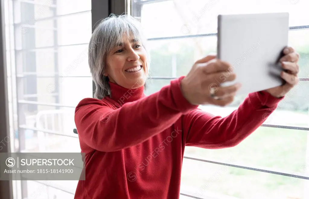 Smiling woman taking selfie through digital tablet by window at home