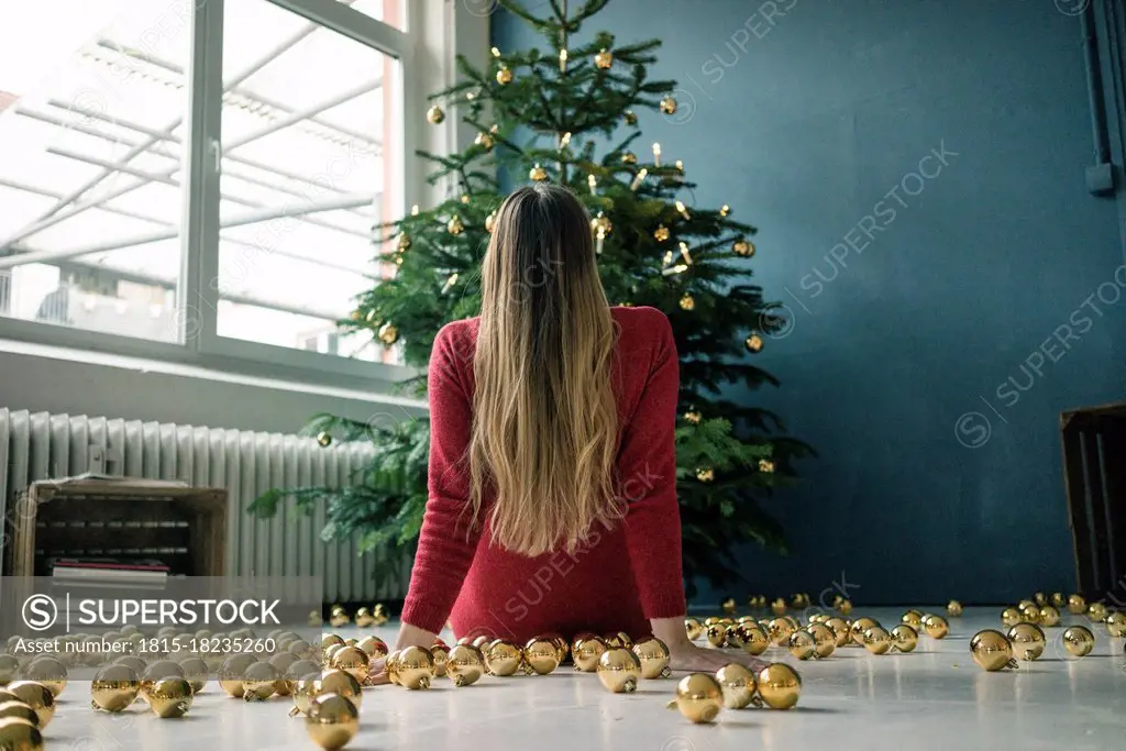 Back view of woman sitting on the floor with many golden Christmas baubles looking at Christmas tree