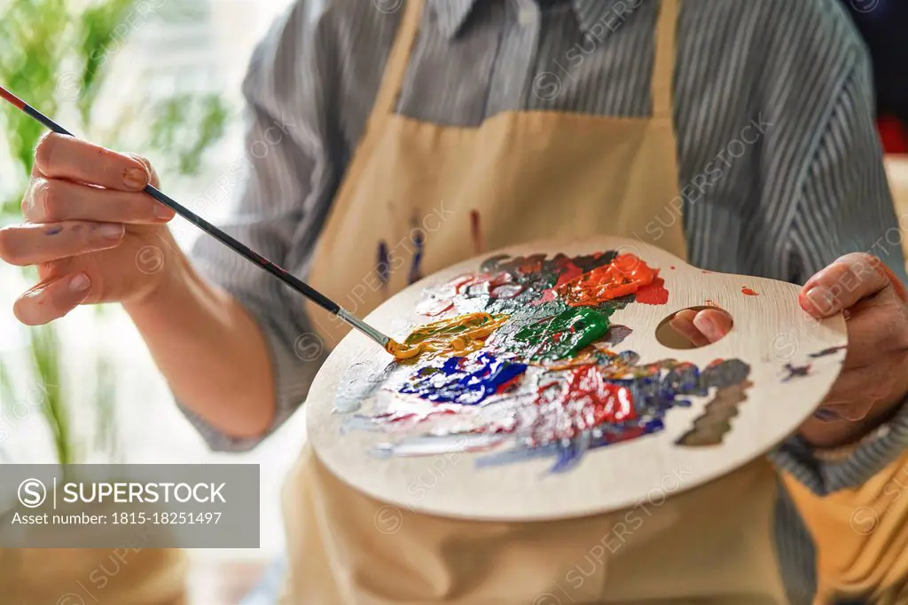 Woman holding palette while mixing colors through paintbrush at home