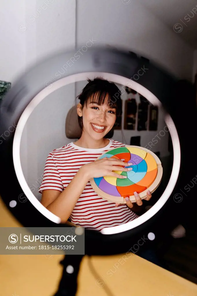 Smiling female influencer seen through ring light while holding multi colored beauty product