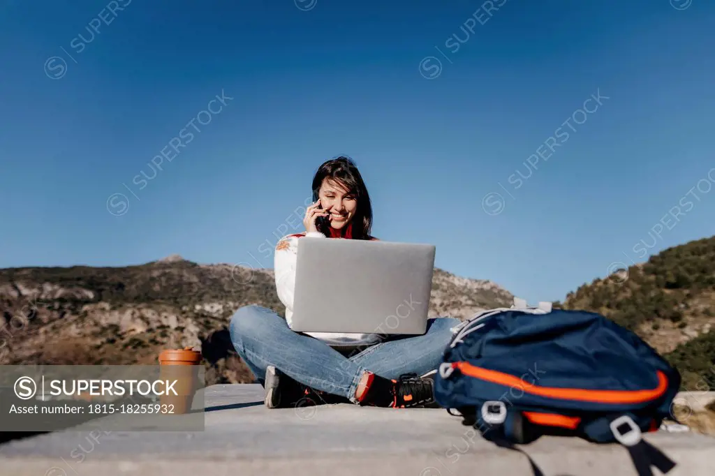 Smiling woman talking on phone while sitting on concrete at mountain