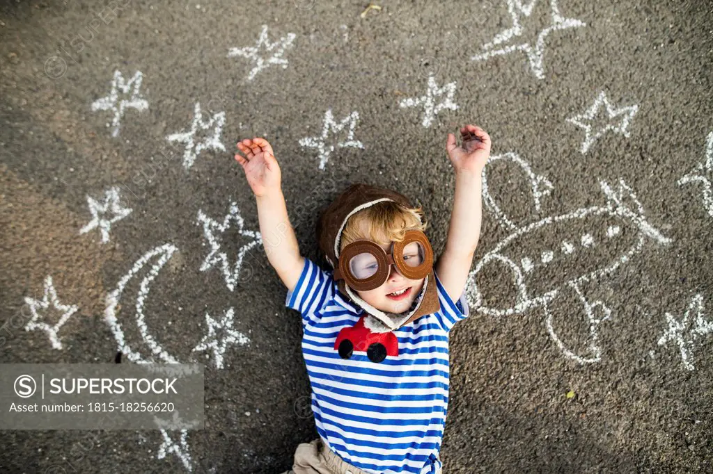 Portrait of smiling toddler wearing pilot hat and goggles lying on asphalt painted with airplane, moon and stars