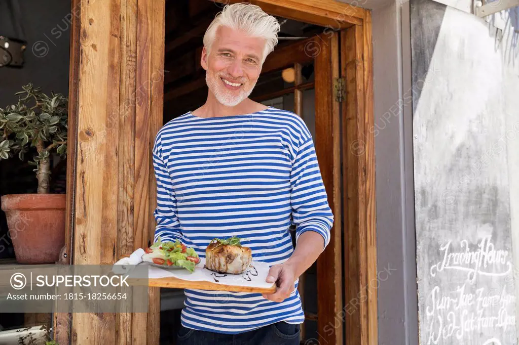 Mature man standing in front of his restaurant, serving a dish