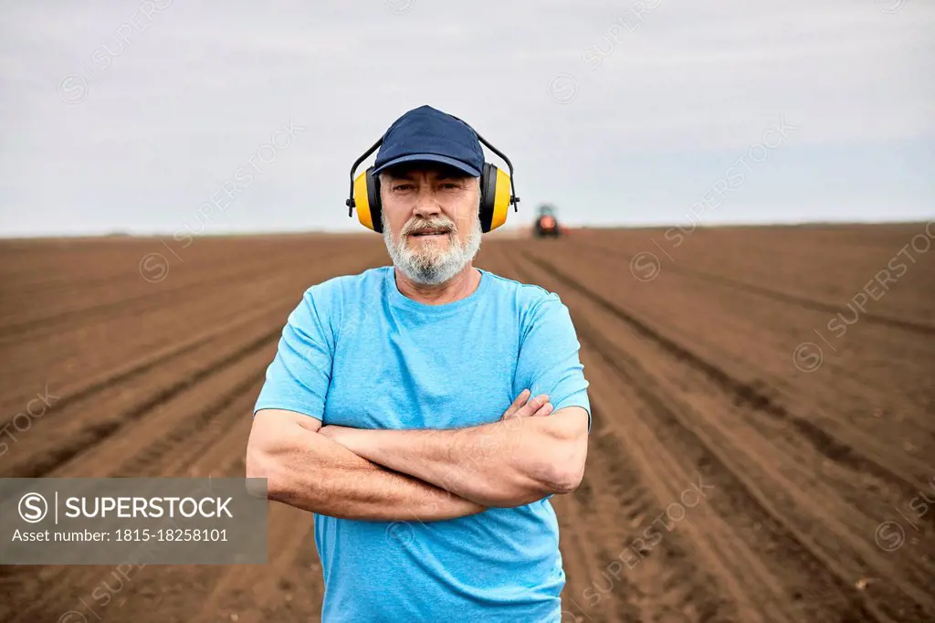 Male farmer wearing ear protectors while standing with arms crossed on agricultural field