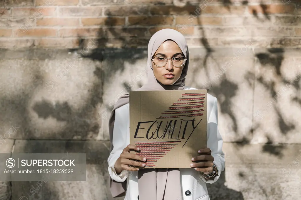 Young woman holding placard with equality text in front of wall on sunny day