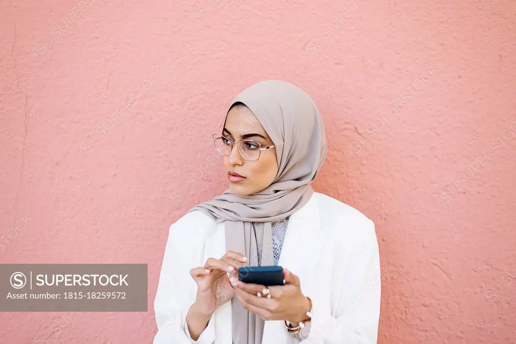 Beautiful woman holding smart phone in front of pink wall looking away