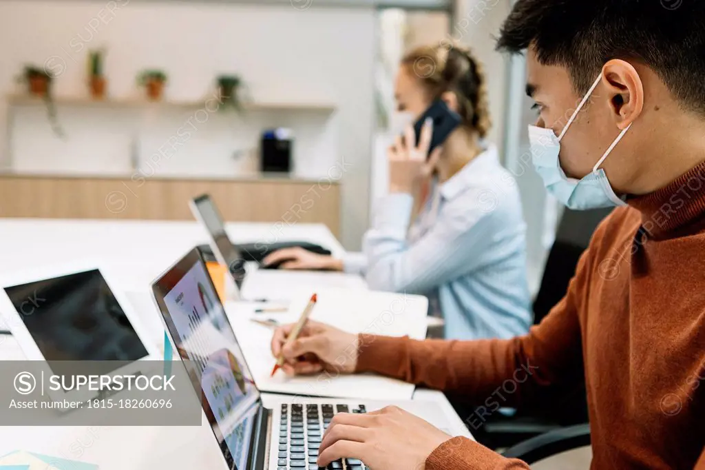 Businessman wearing protective face mask working on laptop with female colleague in background at office