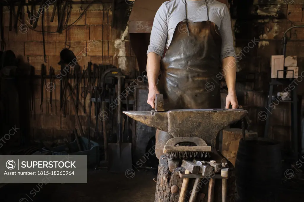 Male blacksmith standing with hammer by anvil at shop