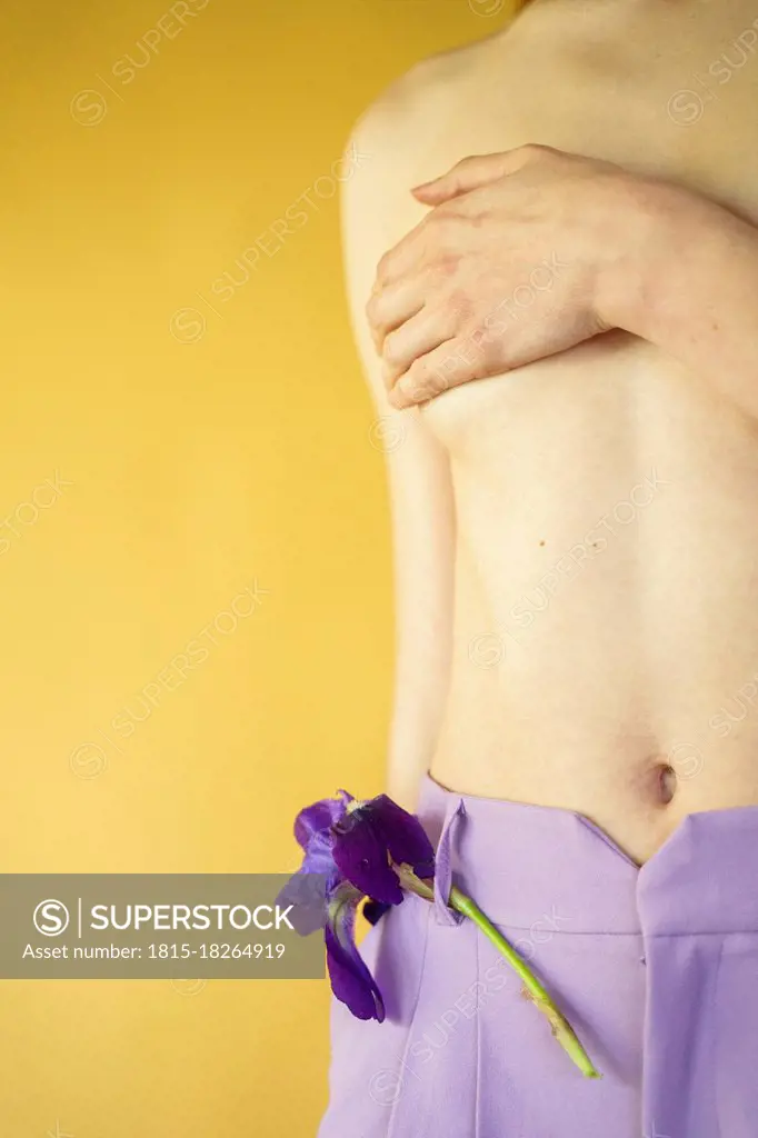 Shirtless woman standing with purple lily flower in pants covering breast in front of yellow wall