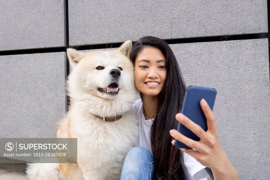 Hipster woman taking selfie with Akita dog through mobile phone by wall