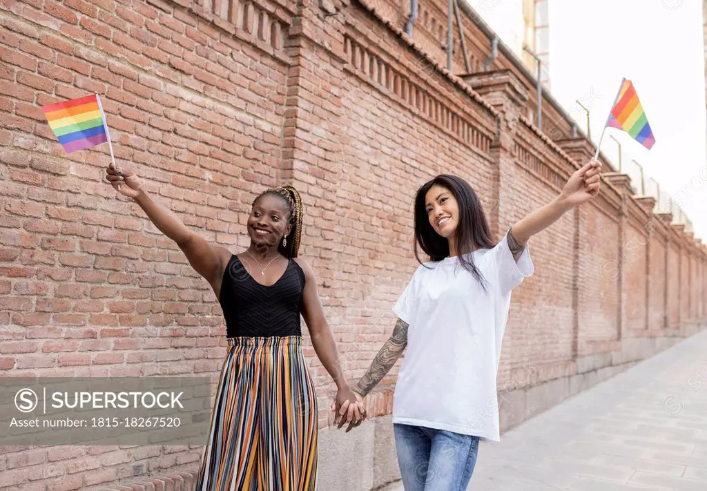 Multi-ethnic lesbian couple holding flag while standing on footpath near wall