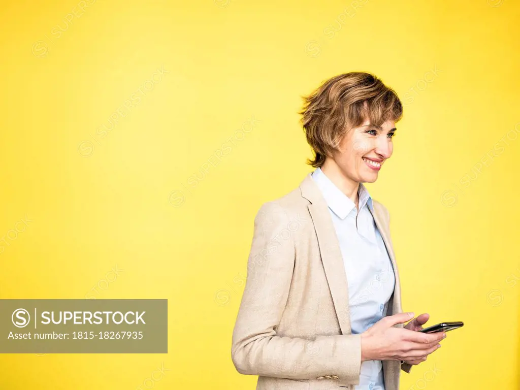 Smiling businesswoman holding mobile phone by yellow background