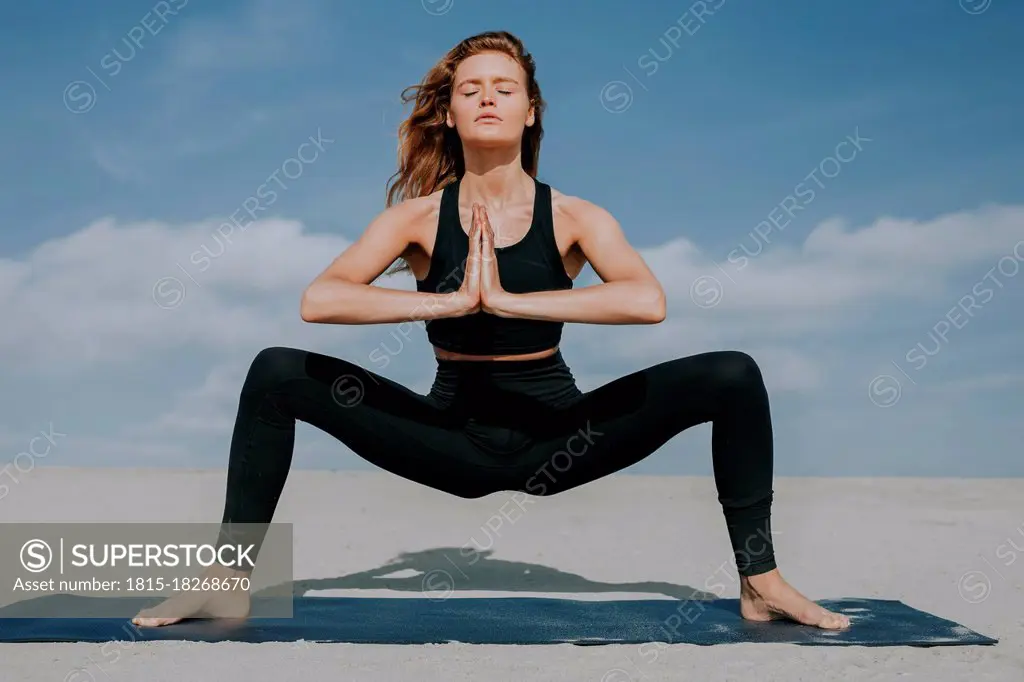 Young woman with hands clasped in squatting position on exercise mat at beach