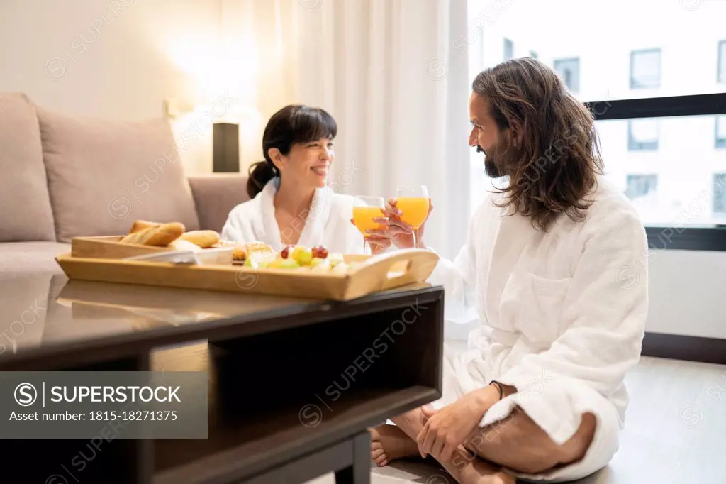 Mature couple toasting glasses of juice while sitting on floor in hotel suite