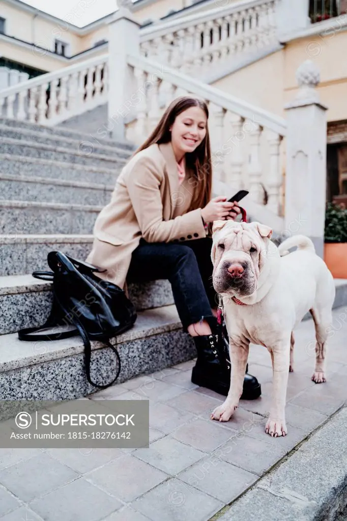 Smiling woman with mobile phone sitting by purebred dog on steps