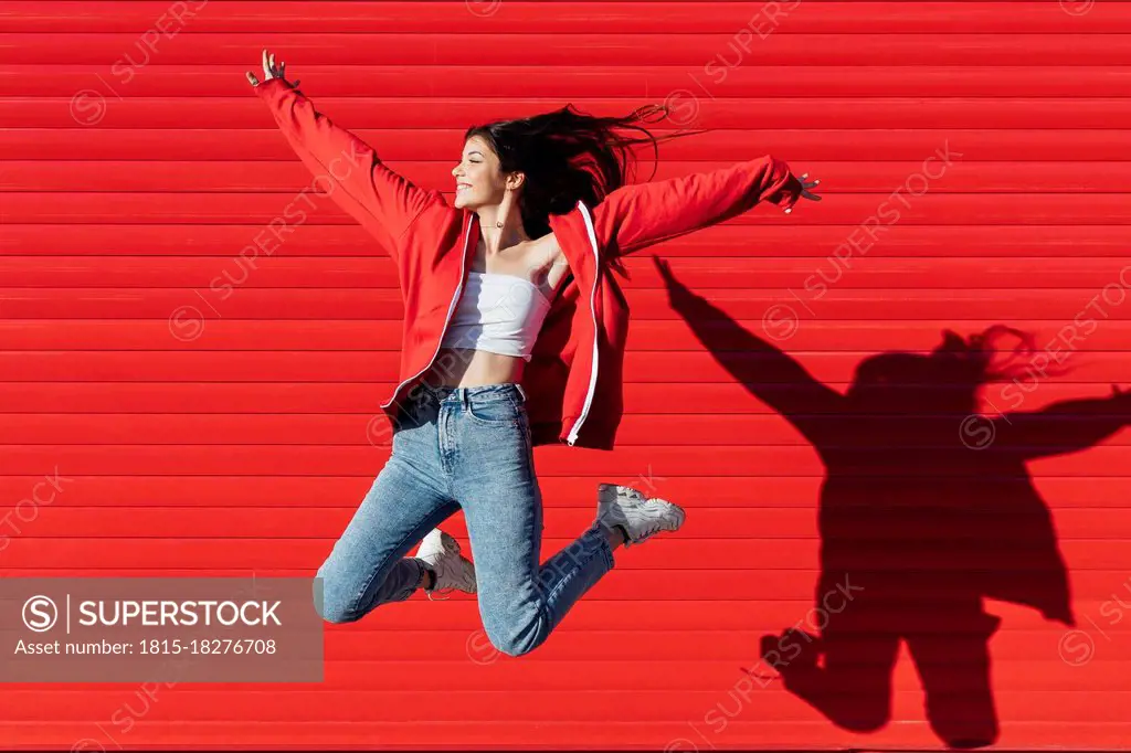 Smiling teenage girl with arms outstretched jumping in front of red wall