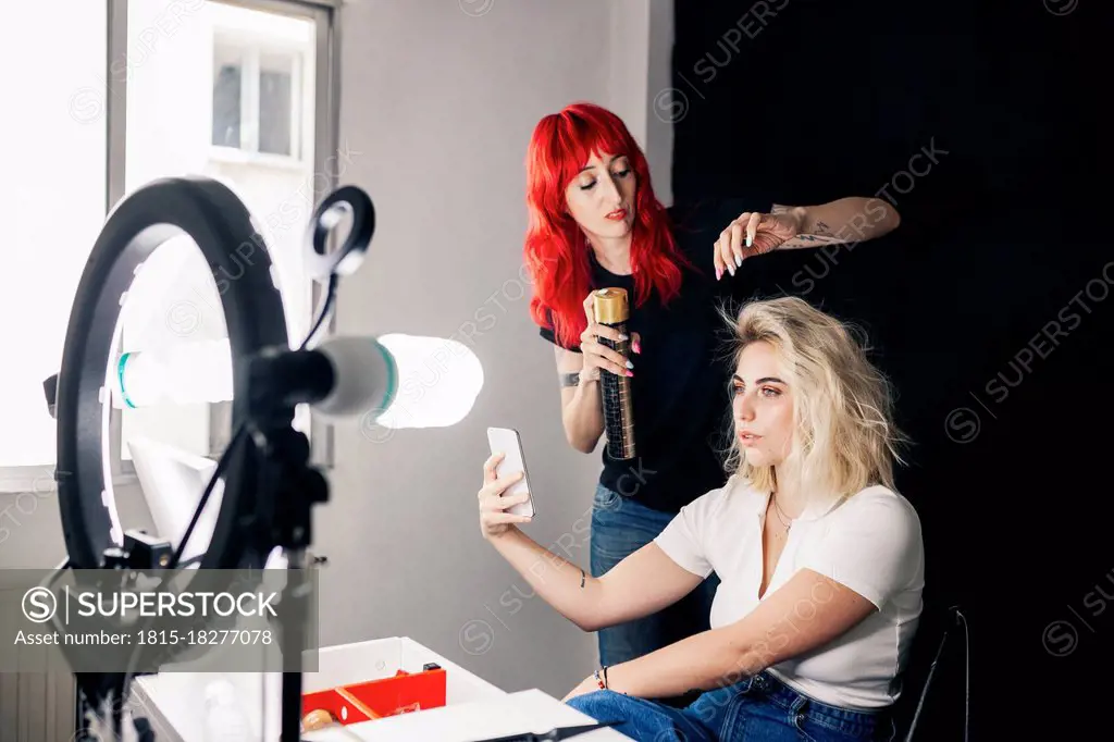 Female stylist applying spray on model's hair taking selfie through mobile phone in studio