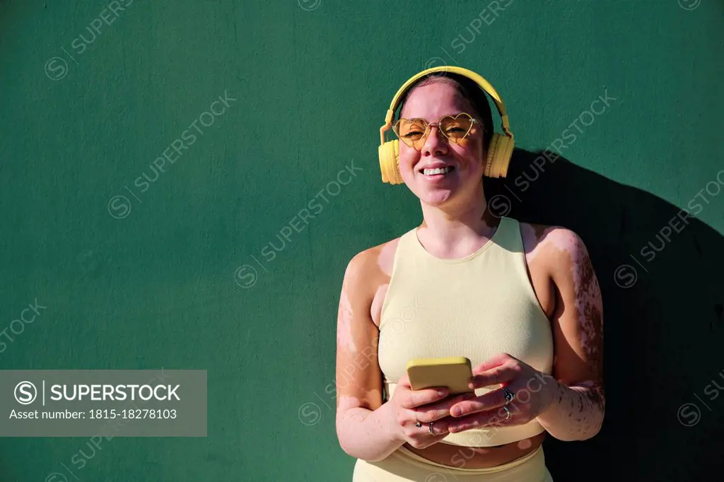 Smiling woman with eyeglasses holding smart phone by green wall