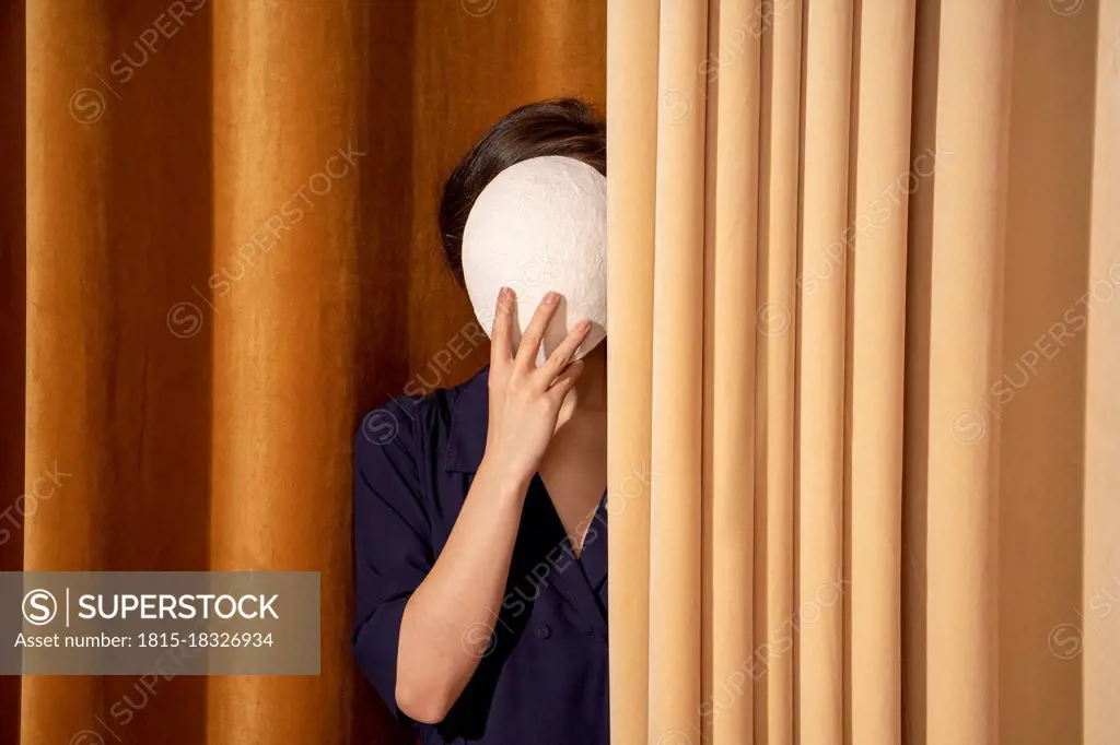 Woman covering face with white mask by curtain