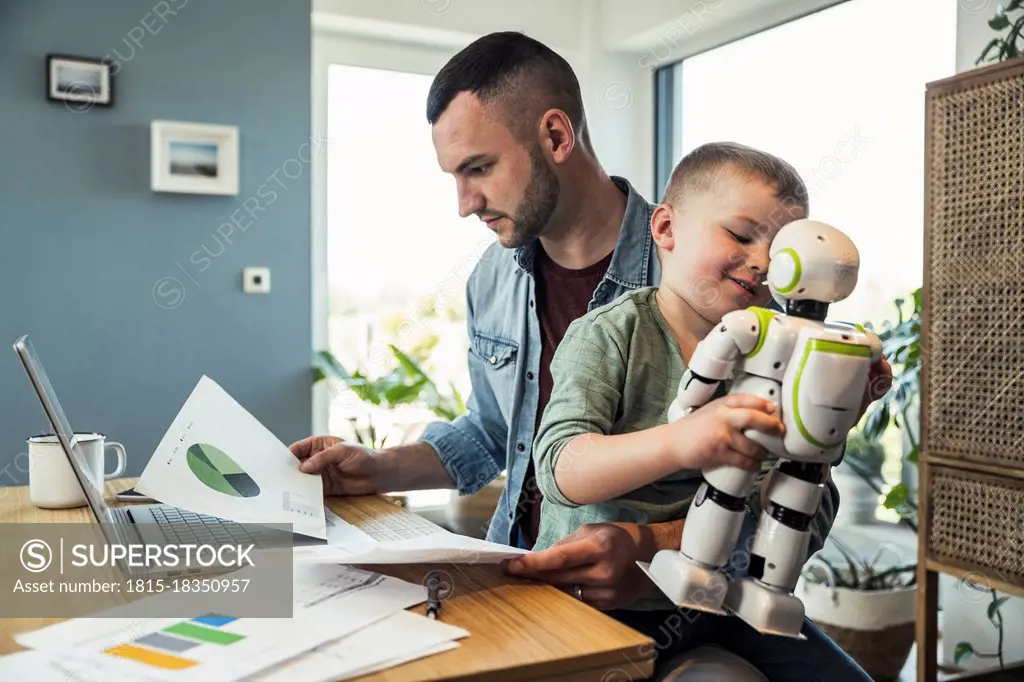 Boy playing with robot sitting while businessman analyzing graph at home