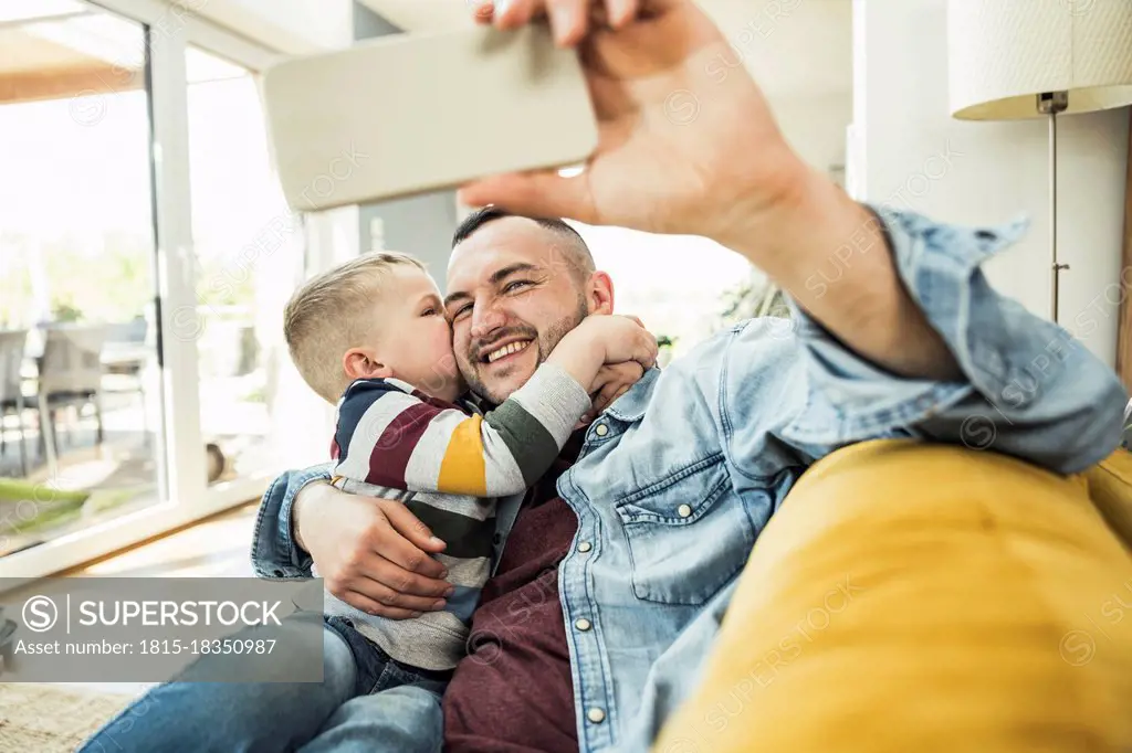 Son kissing on cheek while father taking selfie selfie through mobile phone on living room